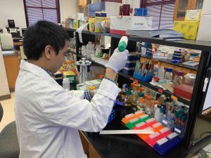 Person in a white lab coat handling a small container at a cluttered laboratory workbench.